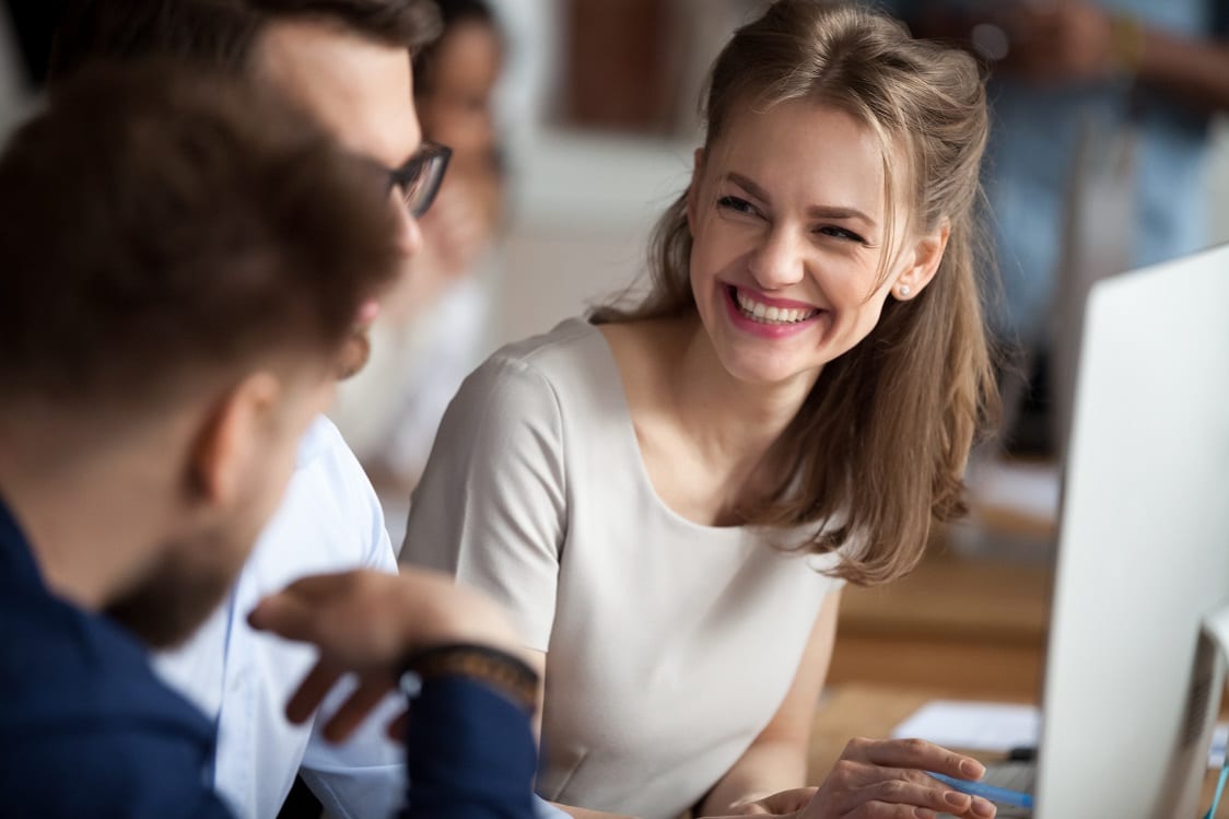 Smiling young woman talking with colleagues at shared workplace Smiling happy young woman talking with male colleagues at shared workplace, worker laughing at funny joke, reading funny news, excited business success, employees having break, pause